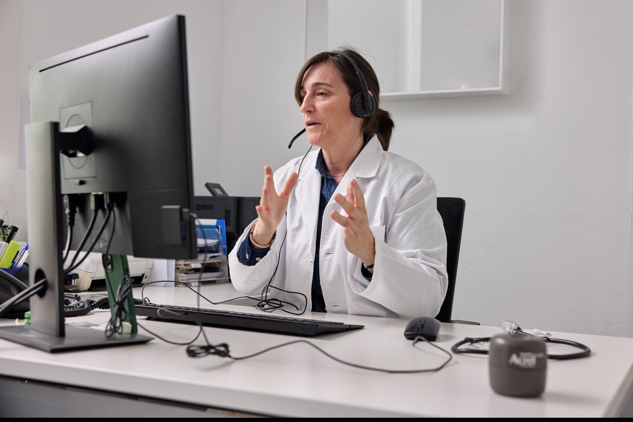 A woman wearing a lab coat is seated in front of a computer, using a headset.